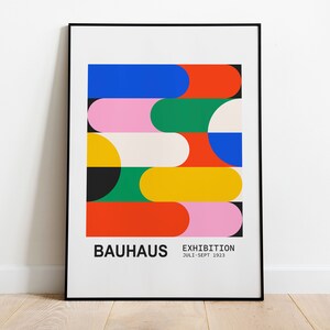 May include: A framed Bauhaus exhibition poster. The artwork features colorful, geometric shapes in red, blue, pink, green, yellow, and white. The poster includes the word "BAUHAUS" and "EXHIBITION JULI-SEPT 1923" in black text.