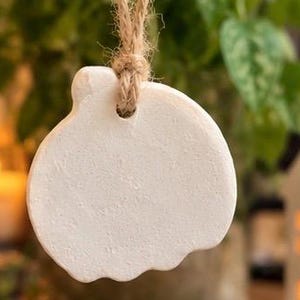 May include: A white, pumpkin-shaped ornament made of a matte, clay-like material. The ornament features a smooth, flat surface with a small hole at the top, through which a piece of natural, light brown jute twine is threaded for hanging purposes.
