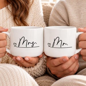 Könnte beinhalten: Zwei weiße Keramiktassen mit schwarzer Schreibschrift. Die linke Tasse zeigt das Wort Mrs. mit einem kleinen Herzsymbol, die rechte Tasse das Wort Mr. mit einem kleinen Herzsymbol. Beide Tassen haben einen einfachen Henkel und eine glatte Oberfläche.
