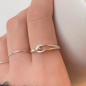 Puede incluir: Primer plano de una mano con tres anillos. El enfoque está en un anillo plateado con un diseño de nudo y una pequeña cuenta dorada. También son visibles otros dos anillos finos de plata. La mano sostiene una taza blanca.