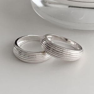 May include: Two silver rings with a ribbed design are shown on a white surface. The rings are next to a clear glass candle holder with the text "Maison Margiela PARIS". The rings have a simple, elegant design.
