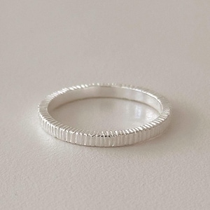 Puede incluir: Un anillo plateado con una superficie texturizada y acanalada. El anillo es un diseño de banda simple, probablemente hecho de plata de ley o un metal similar. La superficie del anillo tiene líneas verticales, creando una textura visual única. El anillo se muestra sobre un fondo neutro.