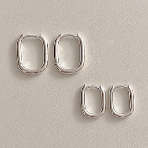 May include: Two pairs of silver oval hoop earrings. The earrings have a smooth, polished finish and a minimalist design. The earrings are displayed on a light grey surface.