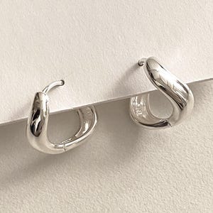 May include: A pair of shiny silver hoop earrings. The earrings have a smooth, curved design and a hinged closure. They are displayed on a white card, showcasing their polished, reflective surface. The earrings are a simple yet elegant accessory.