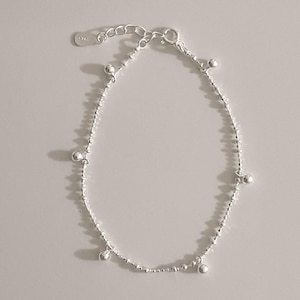 May include: A delicate silver bracelet with small, round charms evenly spaced along the chain. The bracelet has a simple design and a secure clasp. The jewelry is displayed on a light-coloured surface, highlighting its reflective qualities.