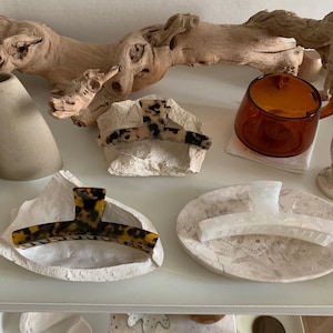 Puede incluir: Tres pinzas para el pelo sobre diferentes bases de piedra. Las pinzas tienen todas la forma de un semicírculo con una parte superior plana. La primera pinza es de carey marrón y negro, la segunda es blanca e iridiscente, y la tercera es de carey marrón y negro.