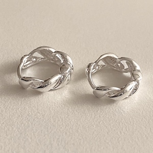 May include: A pair of silver hoop earrings. Each earring has a braided design, with a smooth, reflective surface. The earrings are circular and appear to be made of metal. The background is a neutral, off-white colour.