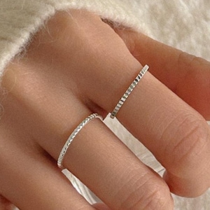 Puede incluir: Dos delicados anillos de plata, cada uno adornado con pequeños adornos brillantes, se llevan en los dedos. Los anillos destacan sobre un fondo suave y neutro, resaltando su simple elegancia. Los anillos son una joya clásica.