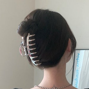 Puede incluir: Una pinza para el pelo de garra plateada sujetando el cabello castaño oscuro de una mujer en un moño. La pinza tiene un diseño único con múltiples dientes. La mujer lleva una camisa negra y un collar de cadena plateada.