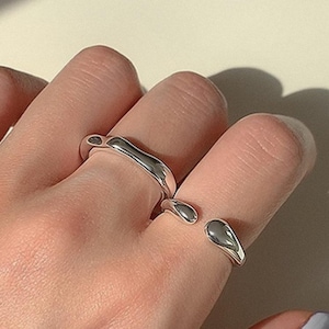 Op de afbeelding: Een zilveren ring met een uniek, abstract ontwerp. De ring is gemaakt van twee delen die aan elkaar zijn verbonden. De ring wordt gedragen aan de middelvinger van een hand.