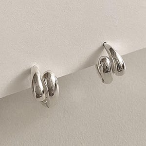 May include: A pair of silver hoop earrings with a distinctive double-loop design. The earrings are presented against a white backdrop, highlighting their polished, reflective surface. The minimalist style of the earrings makes them suitable for various outfits.