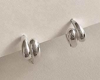 May include: A pair of silver-colored hoop earrings with a unique, double-loop design. The earrings are displayed on a white background, showcasing their polished, reflective surface. The minimalist style of the earrings makes them suitable for various outfits.