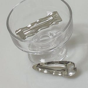 Può includere: Due fermagli per capelli argentati. Uno è un fermaglio rettangolare ondulato con tre creste. L'altro è un fermaglio più piccolo e curvo.