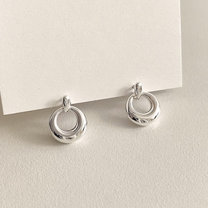Pode incluir: Um par de brincos prateados brilhantes. Cada brinco apresenta um pequeno topo arredondado ligado a um design circular maior. Os brincos têm uma superfície lisa e reflexiva e são exibidos em um fundo branco.
