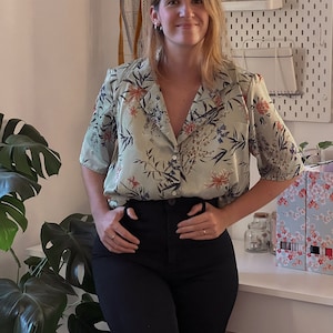 Può includere: Una donna indossa una camicia verde con bottoni e stampa floreale e pantaloni neri a vita alta. È in piedi davanti a una bacheca bianca con vari accessori per la cucito.