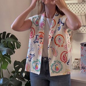 Könnte beinhalten: Eine weiße ärmellose Weste mit einem bunten Paisley-Muster. Die Weste hat einen Schalkragen und wird über einem weißen T-Shirt und einer blauen Jeans getragen.