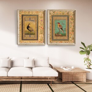 May include: Two framed paintings with ornate gold frames. The paintings feature detailed illustrations of birds on a light blue background with intricate floral borders.