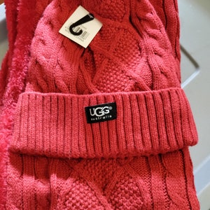 May include: Red knitted hat and scarf set with a pom pom on top of the hat. The hat has a UGG Australia label.