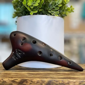 May include: A brown and black ocarina with six finger holes. The ocarina is sitting on a wooden surface.