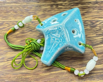 Puede incluir: Una ocarina de cerámica azul con un cordón verde y naranja y cuentas blancas. La ocarina tiene un esmalte craquelado y tiene forma de triángulo.