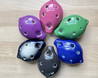 May include: Six ceramic ocarinas in different colors, including purple, green, blue, black, and white. The ocarinas are arranged in a circle on a wooden surface.