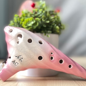 May include: A pink and white ceramic ocarina with a crackled glaze finish. It has eight finger holes and a small hole on the side. The ocarina has Chinese characters and the letters "AC" inscribed on the side.