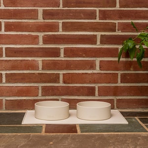 Pu&ograve; includere: Due ciotole in ceramica bianca per animali domestici su un tappetino rettangolare bianco. Le ciotole sono su una superficie piastrellata davanti a un muro di mattoni.