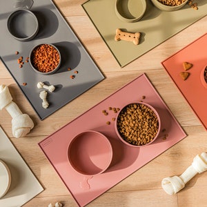 Pu&ograve; includere: Quattro tappetini in silicone per cibo per animali domestici in diversi colori, ognuno con due ciotole. I tappetini sono su una superficie di legno. Le ciotole sono piene di cibo secco per animali domestici. Ci sono anche ossa per cani sui tappetini.