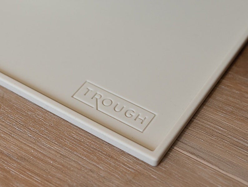 Pu&ograve; includere: Primo piano di un tappetino in silicone beige chiaro con la parola "TROUGH" in rilievo in un rettangolo sulla superficie.