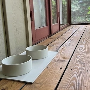 Pu&ograve; includere: Due ciotole per animali domestici in ceramica grigia chiaro su un tappetino rettangolare bianco. Le ciotole sono su un pavimento in legno.