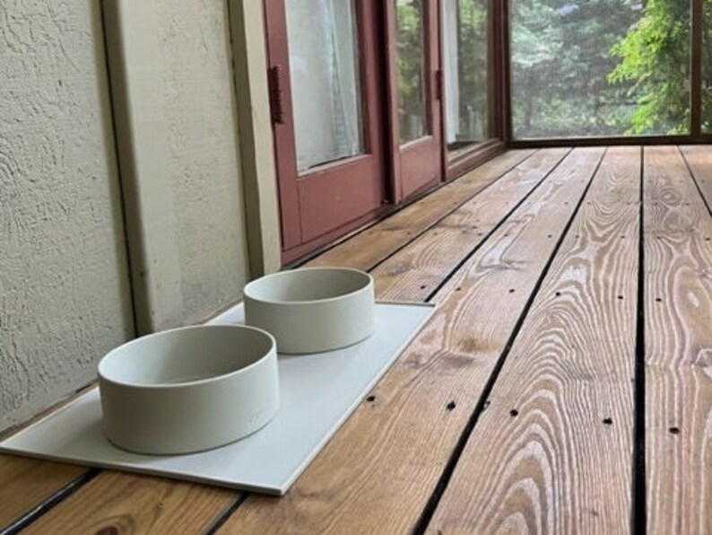 Pu&ograve; includere: Due ciotole per animali domestici in ceramica grigia chiaro su un tappetino rettangolare bianco. Le ciotole sono su un pavimento in legno.