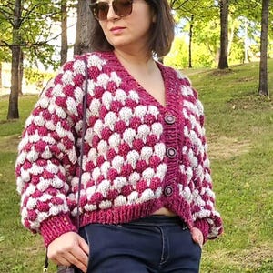 Könnte beinhalten: Burgunderfarbene und weiße Strickjacke mit Blasenmuster. Der V-Ausschnitt-Pullover hat eine Knopfleiste vorne und lange Ärmel. Die Person trägt eine dunkelblaue Jeans und eine Sonnenbrille. Eine kleine lila Handtasche hängt an der Schulter.