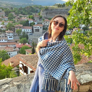 May include: A woman wearing sunglasses and a blue and white patterned shawl with fringe. The shawl is draped over a blue outfit. The background features a scenic view of a town with red-tiled roofs and green trees.