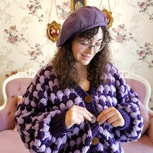 Könnte beinhalten: Ein lila und lavendelfarbener Strickcardigan mit einem karierten Muster und Holzknöpfen. Der Cardigan ist kurz und hat einen V-Ausschnitt. Das Model trägt eine lilafarbene Baskenmütze und eine Brille.