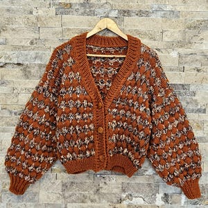 Könnte beinhalten: Ein brauner und weißer Strickcardigan mit Knopfleiste und langen Ärmeln. Der Cardigan hat ein strukturiertes Muster und einen V-Ausschnitt.