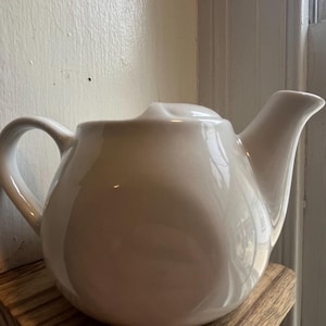 May include: A white ceramic teapot with a rounded body and a spout. The teapot is sitting on a wooden shelf.
