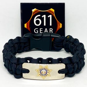 May include: Black paracord bracelet with a black plastic clasp and a silver-coloured metal plate. The plate features a security officer badge with the words "SECURITY OFFICER" and a star design. The background features a black sign with the text "611 GEAR".