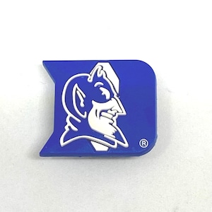 Puede incluir: Un pin con el logotipo de la Universidad de Duke en azul y blanco. El pin presenta la mascota del diablo de la universidad en blanco sobre un fondo azul. El pin tiene la forma de una letra "D" estilizada con un pequeño símbolo "R".