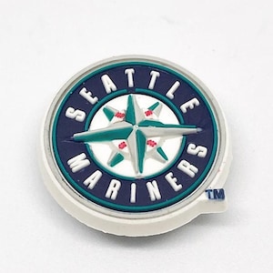 May include: A round, white, blue, and teal Seattle Mariners logo. The logo features a compass rose design in teal and red, surrounded by a navy blue ring with the words "SEATTLE MARINERS" in white. A small "TM" is visible.