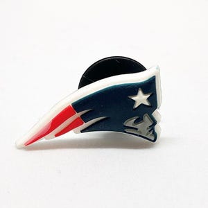 May include: A colorful shoe charm featuring the New England Patriots logo. The charm has a white, red, and blue color scheme, with a star and a profile of a man's face. The charm is designed to attach to footwear.