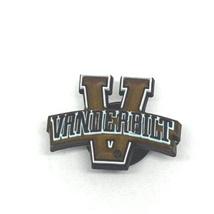 May include: A brown and white Vanderbilt University logo pin. The pin features a stylized letter "V" above the word "VANDERBILT" in white lettering. The pin is set against a white background.