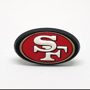 Puede incluir: Un dije ovalado con el logotipo de los 49ers de San Francisco. El logotipo tiene un fondo rojo con un borde dorado y un emblema "SF" blanco. El dije tiene un borde negro.