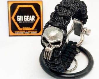 May include: Black paracord bracelet with a silver skull charm. The bracelet features a metal clasp and a black ring. The logo for 611 Gear is visible in the background.