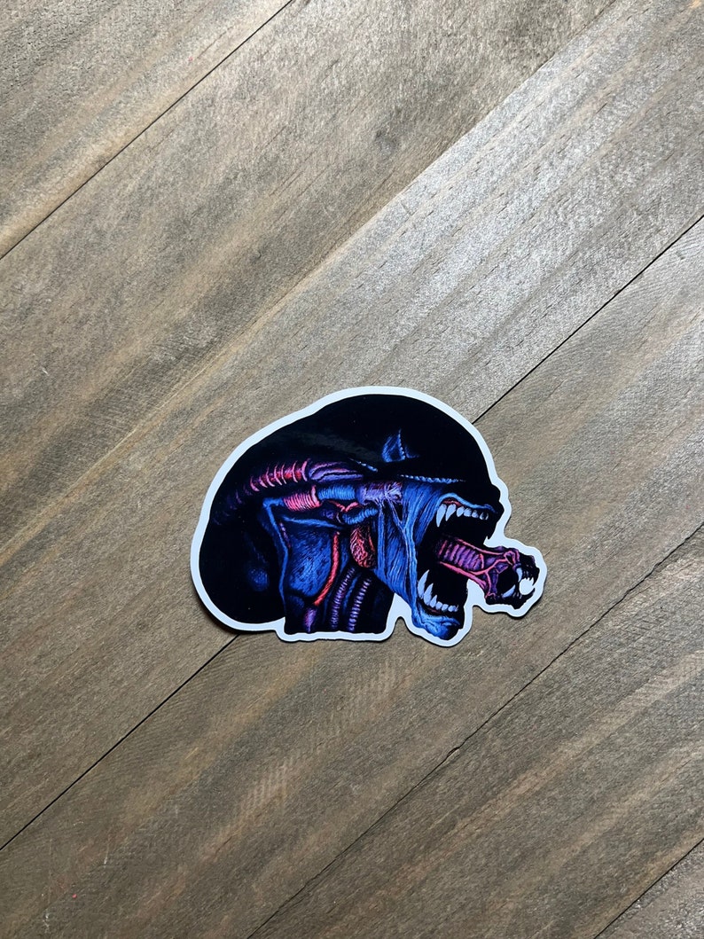 May include: A black and blue alien creature sticker with a white outline. The alien has a large mouth with sharp teeth and a long, thin tongue.