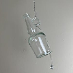 May include: A clear glass bottle with a silver chain attached to the top. The bottle is hanging upside down and the chain is attached to a silver charm.
