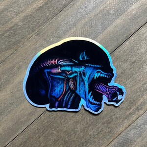 May include: A holographic sticker featuring a blue and purple alien creature with a large mouth and sharp teeth. The alien is shown with its mouth open, revealing a long, thin tongue.