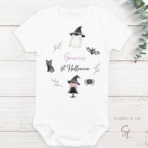 Może przedstawiać: Biały śpioszek dla niemowlęcia z czarnym kotem, czarownicą, duchem, nietoperzem i pająkiem. Śpioszek ma napis "Gracie's 1st Halloween" w fioletowym kolorze.