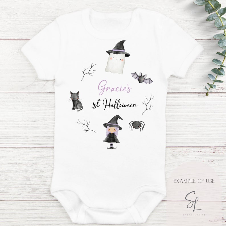Może przedstawiać: Biały śpioszek dla niemowlęcia z czarnym kotem, czarownicą, duchem, nietoperzem i pająkiem. Śpioszek ma napis "Gracie's 1st Halloween" w fioletowym kolorze.