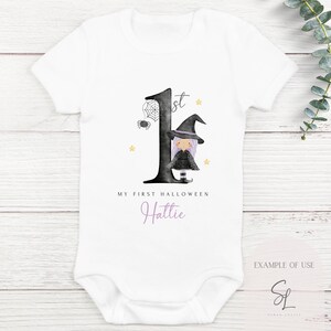 Może przedstawiać: Biały śpioszek dla niemowlęcia z czarną jedynką i ilustracją czarownicy. Na śpioszku nadrukowany jest tekst "My First Halloween Hattie".