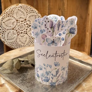 May include: A cylindrical container with a floral design and the word "Seelentröster" printed on it. The container is filled with floral cutouts and sits on a silver tray, which is placed on a wooden tray.